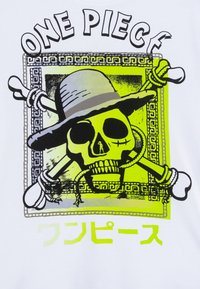 Balta T-kreklā ir grafiskais dizains ar skeletu, kas valkā cepuri un kauliem krustā, akcentēts dzeltenā krāsā ar melniem elementiem un japāņu tekstu.