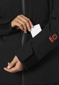Černá bunda s ozdobným zipem a manžetami na suchý zip. Ruka vkládá bílou kartu do kapsy bundy. Viditelné oranžové logo.