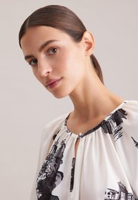 Blusa bianca con stampa del paesaggio urbano nera, scollo arricciato e dettaglio con apertura a chiave. Tessuto morbido con una consistenza liscia e vestibilità rilassata.