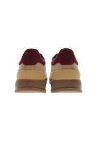 Hnědé tenisky s hnědou gumovou podrážkou, které mají červený a námořnický látkový límec a branding. Design je zvýrazněn zúženými liniemi a polstrovaným jazýčkem.