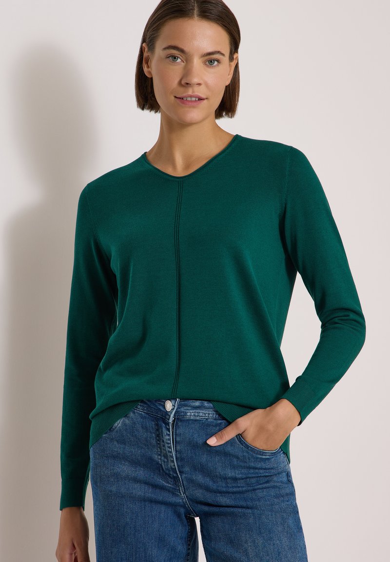Femme portant un pull à manches longues vert foncé et un jean bleu, une main dans la poche, sur un fond clair uni.