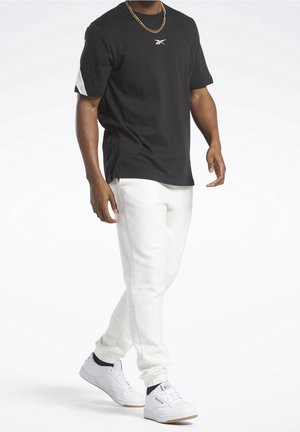 Camiseta de algodón negra con detalles blancos y logotipo, combinada con joggers blancos y zapatillas blancas. Cadena de oro al cuello.