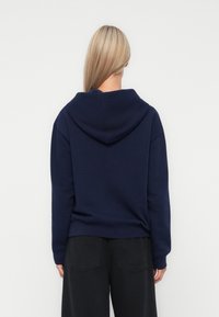Marinblå sweatshirt med huva, mjuk textur, lös passform och ribbade ärmmuddar. Här tillsammans med svarta vida byxor, vilket ger en avslappnad stil.