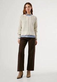 Cream knitted sweater with openwork detailing, layered over a blue-striped shirt. Paired with brown wide-leg trousers and beige heeled boots.