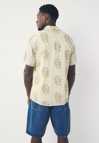 Camicia a maniche corte color beige chiaro con righe verticali e motivi floreali in rosa, blu e verde. Abbinata a pantaloncini di jeans.