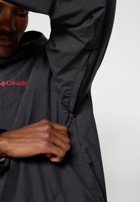 Μαύρο αδιάβροχο μπουφάν με φερμουάρ στην αριστερή μανσέτα, με κόκκινο λογότυπο Columbia μπροστά. Λείο υλικό, μοντέρνος σχεδιασμός.