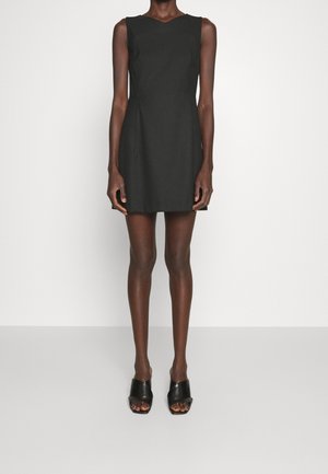 Modelo delgado vistiendo un mini vestido negro sin mangas y sandalias de tacón abiertas negras, de pie contra un fondo blanco liso.