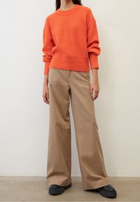 Oranssi ribbineulottu pusero, jossa leveät hihat, yhdistettynä beigeihin leveälahkeisiin housuihin ja mustiin korotettuihin kenkiin, neutraalia taustaa vasten.