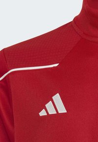 Haut de sport rouge à manches longues avec un col montant, en tissu texturé, accents blancs le long des épaules et un logo blanc comportant trois bandes.