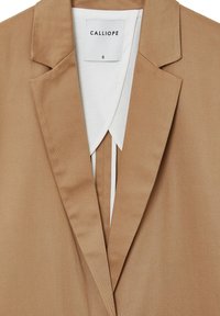 Blazer beige avec un col cranté, un design structuré et une texture lisse. La doublure intérieure blanche présente un pli vertical au centre.