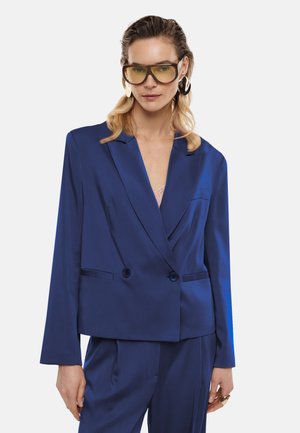 Donna che indossa occhiali oversize con lenti gialle e grandi orecchini, vestita con un blazer blu lucido e pantaloni coordinati.