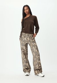 Maglione in maglia marrone a spalla unica abbinato a jeans a vita alta con motivo camouflage grigio e beige. Il modello indossa sneakers bianche.