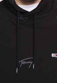 Sudadera negra con capucha ajustable, logo bordado "Tommy" en gris y "JEANS" impreso en los extremos de los cordones.