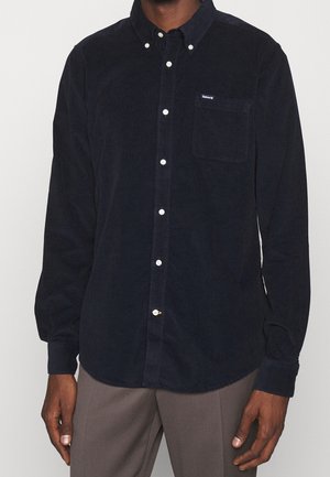 Homme portant une chemise noire en velours côtelé à manches longues, boutonnée avec une poche poitrine, et un pantalon gris sur un fond uni.