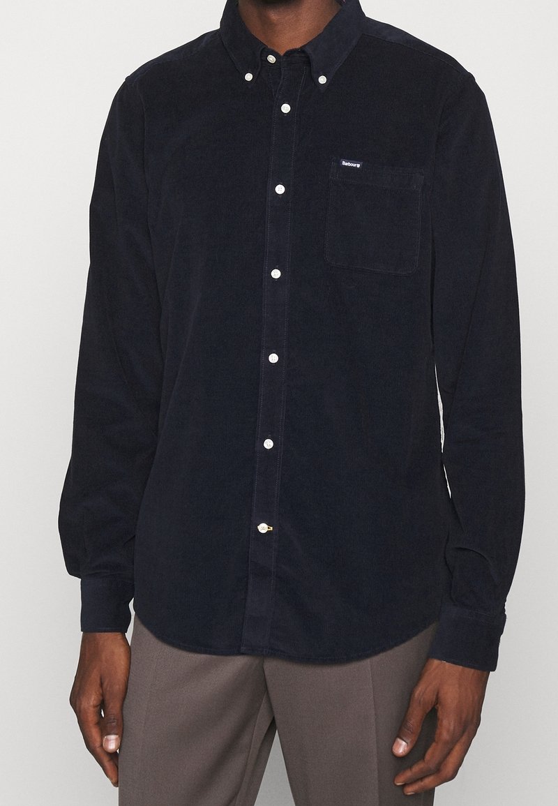 Homme portant une chemise noire en velours côtelé à manches longues, boutonnée avec une poche poitrine, et un pantalon gris sur un fond uni.