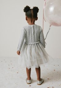 Grijze gebreide cardigan met kabelpatroon, gecombineerd met een witte tule jurk met pailletten. Zilveren glitterschoenen en een ballon accentueren de outfit.