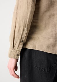 Camisa de lino beige con mangas enrolladas y detalle de botones, combinada con pantalones de mezclilla negra oscura. Muestra contraste de textura y material entre las telas.