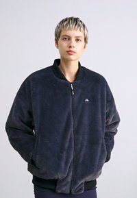 Jachetă din flanel gri închis, cu guler înalt, fermoar frontal și manșete elastice. Dispune de un logo mic pe pieptul stâng. Textură moale.