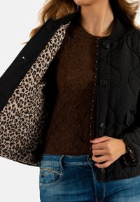 Zwarte gewatteerde jas met een voering van luipaardprint, gedragen over een bruine gebreide trui. Zichtbare knopen en een jeans tailleband zijn aanwezig.
