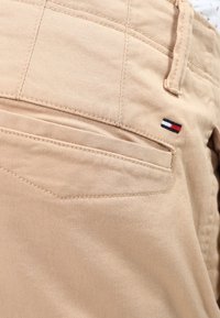 Beige cotton pants featuring a back pocket with a small embroidered logo in red, white, and blue. Smooth texture with fine stitching details.