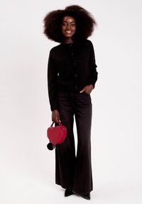 Cardigan en maille noire avec un col rond et des boutons en forme de cœur, associé à un jean flare foncé. Tenue d'un sac à main en forme de cœur rouge avec un pompon.