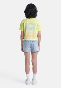 Νεαρός με μαλλιά μέχρι τους ώμους στέκεται με την πλάτη, φορώντας κίτρινη μπλούζα με σχέδιο, ανοιχτόχρωμα τζιν σορτς, λευκές κάλτσες και λευκά αθλητικά παπούτσια.