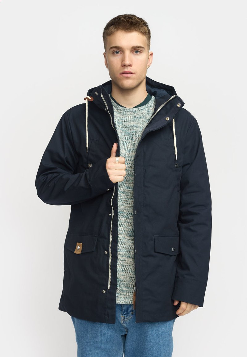 Navy parka jacket with a hood, toggle drawstring, front zip closure, and two snap pockets; layered over a knitted sweater and blue jeans.