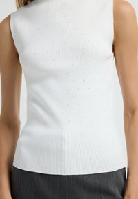 Nahaufnahme einer Person, die ein ärmelloses weißes Rippenoberteil mit kleinen silbernen Punkten und dunkelschwarzen Pants trägt.