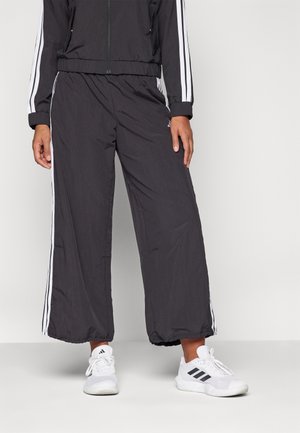 Person, die schwarze, weit geschnittene Jogginghosen mit weißen Seitenstreifen und weißen Adidas-Sneakern trägt und vor einem schlichten hellen Hintergrund steht.