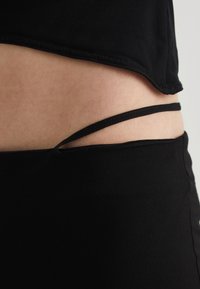 Haut ajusté noir en tissu lisse, avec un ourlet incurvé et de fines bretelles noires visibles au niveau de la taille. La peau est visible en dessous.