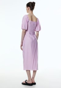 Katoenen jurk in lichtroze met een vierkante rug, pofmouwen en gesmokte details. Middellange ontwerp, gecombineerd met chunky zwarte sandalen.