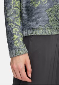 Strickpullover mit einem strukturierten grauen Körper, grünen Blumenmustern und Pailletten. Der Bund ist gerippt und weist filigrane Details auf. Unten ist schwarzer Stoff.