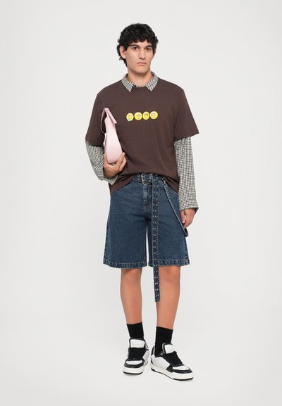 Jeune homme portant un T-shirt emoji marron par-dessus une chemise à manches longues à carreaux, un short en denim avec une ceinture longue, des chaussettes noires, des baskets blanches, tenant un sac rose.