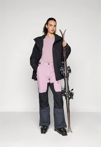 Γυναίκα με ροζ και μαύρο παντελόνι σκι, μαύρο μπουφάν και μπότες σκι, κρατώντας ένα ζευγάρι σκι όρθιο δίπλα της.