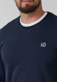 Bearded man wearing a navy blue sweatshirt with a white crew neck shirt underneath, featuring a small "s.O" logo on the chest.