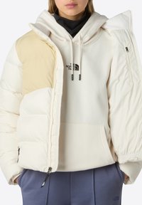 Person klädd i en beige och krämfärgad pufferjacka över en krämfärgad hoodie med The North Face-logotyp, tillsammans med blå byxor.