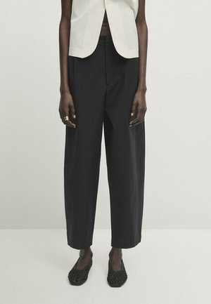Mannequin portant un pantalon noir large raccourci, un haut blanc sans manches, des chaussures plates noires tissées et une bague en or à la main gauche.