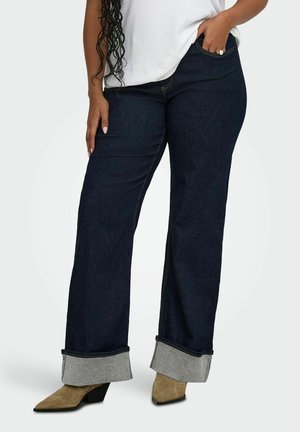 Femme portant un jean large foncé avec des revers pliés, une chemise blanche et des bottines beiges à bout pointu, une main dans la poche.