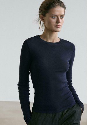 Top aderente a manica lunga in tessuto liscio di colore blu navy con scollo tondo e polsini a coste. Indossato con pantaloni scuri a righe. Design minimalista.