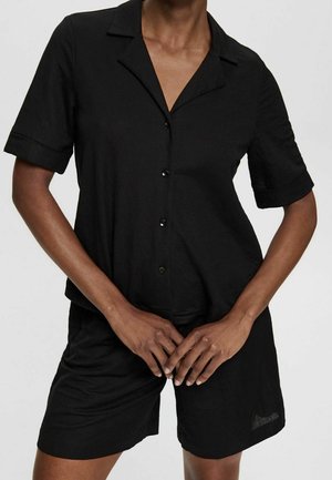 Person iført en kortærmet, sort skjorte med knapper og krave samt matchende sorte shorts, stående mod en ensfarvet baggrund.