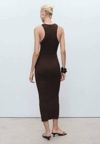 Robe en tricot marron sans manches, au design ajusté, longueur mi-mollet et col haut. Portée avec des accessoires noirs et des sandales.