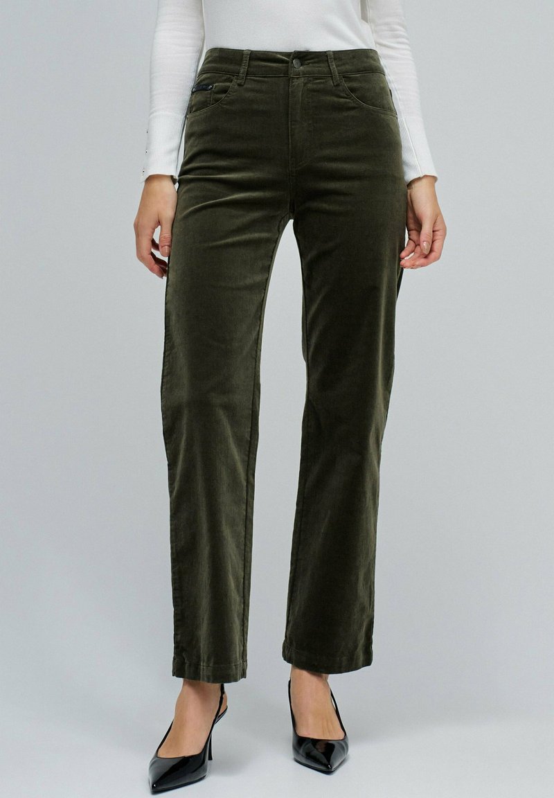 Femme portant un pantalon en velours vert olive foncé taille haute associé à des escarpins noirs à bouts pointus et un haut à manches longues blanc.