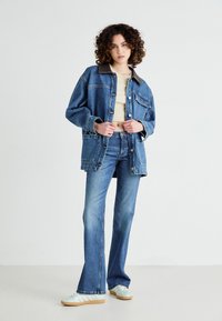 Denim jacket with brown collar, flared denim jeans, and light blue sneakers. The jacket features buttons and pockets, denim in blue tones.