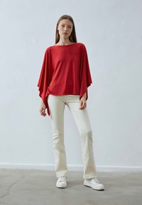 Rotes Batwing-Oberteil aus glattem Stoff, kombiniert mit weißen Schlaghosen und weißen Turnschuhen. Einfaches Design mit lockerer Passform und Drapierung.