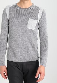 Pull en maille gris avec motif côtelé texturé, épaules contrastantes en gris clair et poche poitrine. Porté avec un pantalon noir.