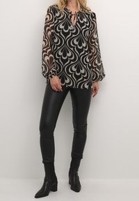 Blouse à motifs noir et beige avec des manches longues, décolleté en trou de serrure et bords froncés, assortie à un legging noir élégant et des bottes pointues.