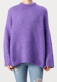 Person klädd i en lös, fluffig lila tröja med långa ärmar och ljusblå ripped jeans, stående mot en enfärgad bakgrund.