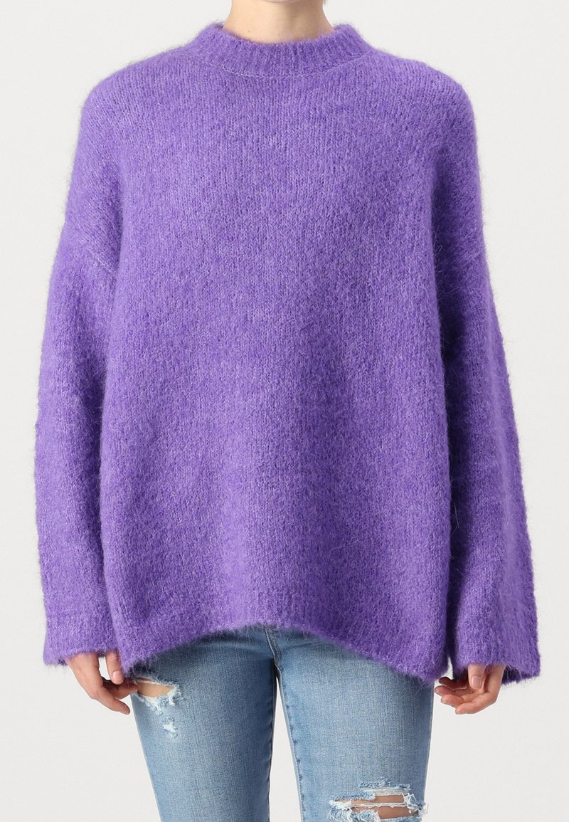 Person klädd i en lös, fluffig lila tröja med långa ärmar och ljusblå ripped jeans, stående mot en enfärgad bakgrund.