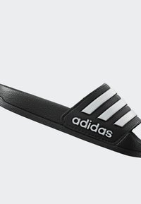 Čierne šľapky s tromi bielymi pruhmi a logom "adidas". Vyrobené z umelého materiálu, s hladkou textúrou a otvoreným dizajnom.