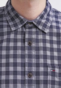 Marineblauw en grijs geblokt overhemd met een klassieke kraag, voorzien van bruine knopen en een klein rood, wit en blauw logo op de zak.